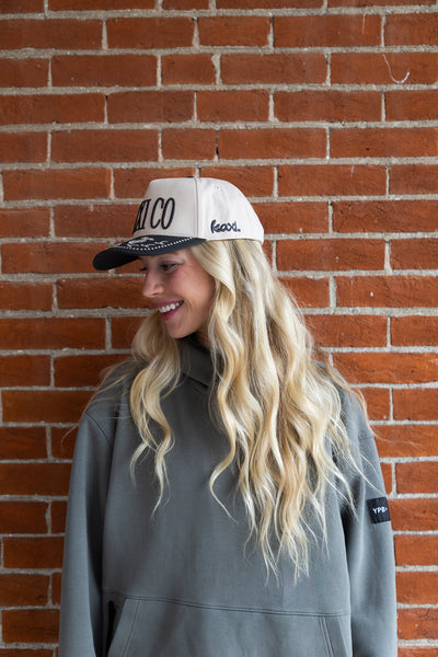 Woman wearing a cap with 'Kaxi Co' logo against a brick wall