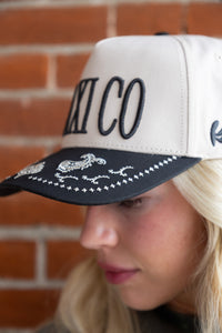 Person wearing a white and black cap with 'MEXICO' and rooster designs against a brick wall.