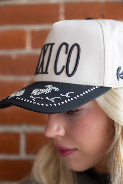 Person wearing a white and black cap with 'MEXICO' and rooster designs against a brick wall.