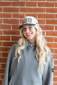 Woman wearing a cap with 'KAXI CO' text against a brick wall