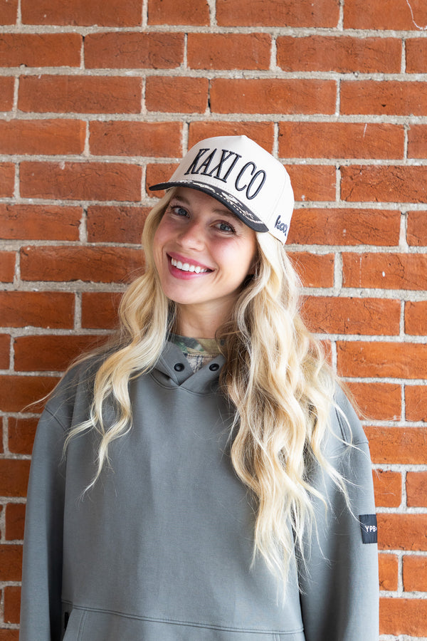 Woman wearing a cap with 'KAXICO' text against a brick wall