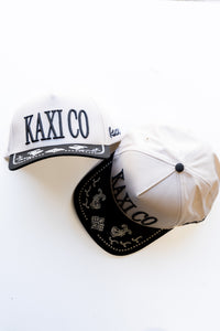 Two white baseball caps with black kaxi co embroidery and embroidered paisley on brim on a white background
