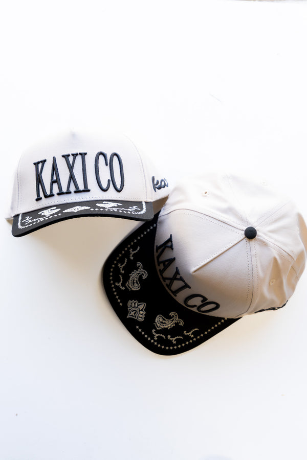 Two white baseball caps with black kaxi co embroidery and embroidered paisley on brim on a white background
