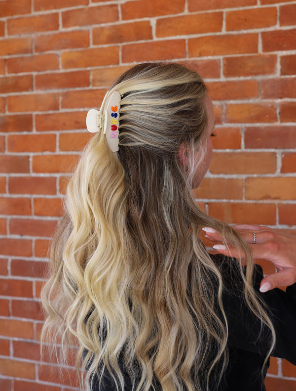 Person with wavy blonde hair styled with a colorful hair clip against a brick wall.