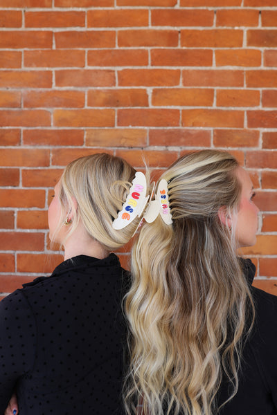 Two women with blonde hair styled with colorful hair clips against a brick wall.