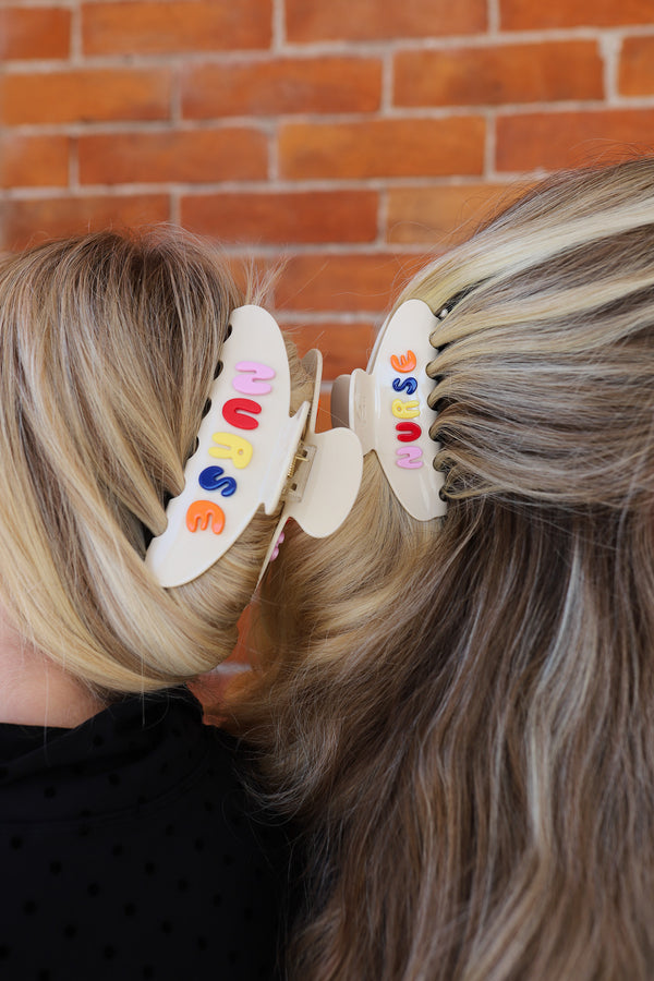 Two hair clips with colorful letters spelling "NURSE" on a person's head against a brick wall.