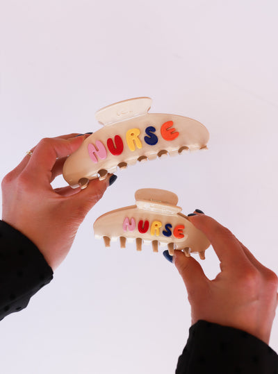 Two hair clips with 'NURSE' letters held by hands against a white background
