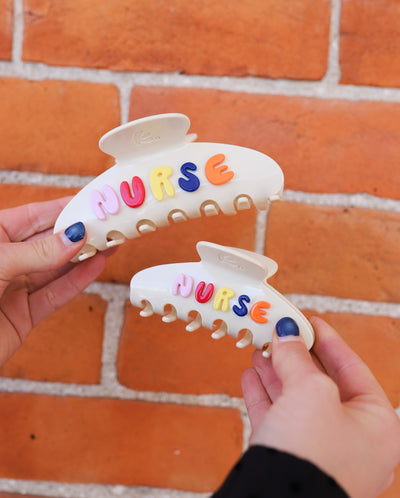 Two hair clips with 'NURSE' in colorful letters held against a brick wall.