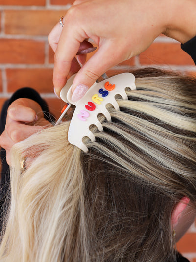 Person putting a cream claw clip with colorful letters spelling "NURSE"  against a brick wall background.