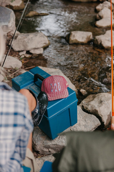 Person fishing by a stream with a trucker hat  on a blue box