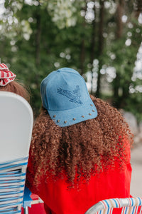 girl with curly hair wearing a backwards baseball hat with an eagle embroidered on it, with studs on the brim