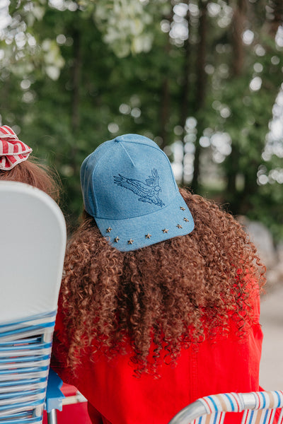 girl with curly hair wearing a backwards baseball hat with an eagle embroidered on it, with studs on the brim