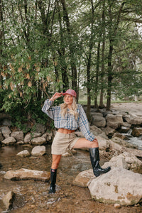 Person standing on rocks by a stream wearing a plaid shirt, shorts, red trucker hat, and rain boots.