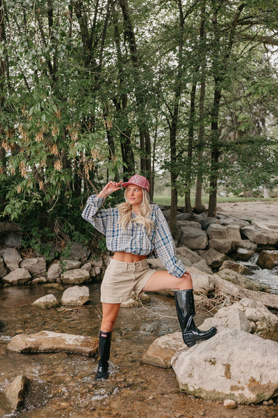 Person standing on rocks by a stream wearing a plaid shirt, shorts, red trucker hat, and rain boots.