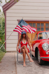 Woman in a red dress, wearing a denim star hat with a tedddy bear embroidered, sitting in front of a American flag