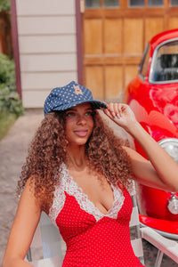 Woman in a red polka dot dress and denim  star baseball cap with a bear embroidered on it  sitting next to a red vintage car.