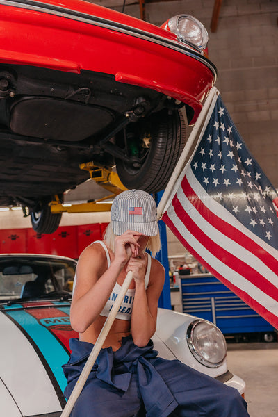 Woman sitting on a car wearing an American flag baseball cap, with an American flag in the background