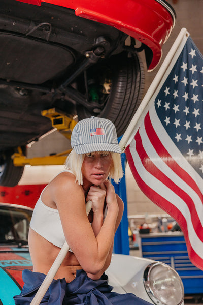 Person with a cap featuring an American flag in front of a car with an open hood, with an American flag in the background.