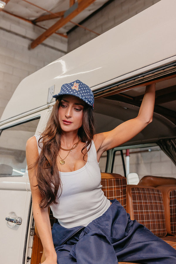 Woman sitting inside a vehicle wearing a white tank top and denim blue star baseball cap with a teddy bear embroidered on the front