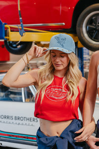 Woman wearing a red tank top and blue denim trucker hat in front of a vintage car and truck.