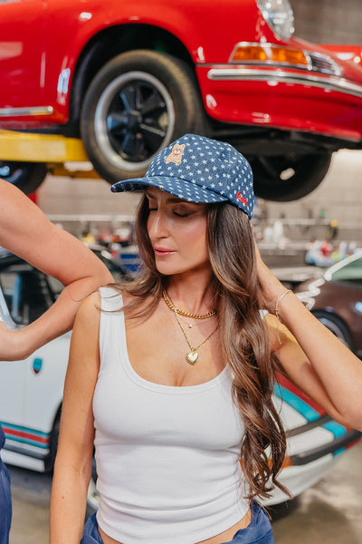 Woman wearing a denim star baseball cap with a teddy bear on it, in a garage with a red car in the background