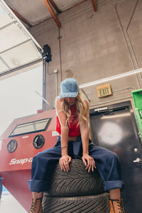 Person sitting on a tire wearing a blue denim baseball cap with an eagle embroidered on it, and star studs on the brim