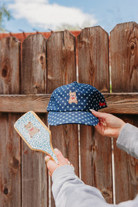Person holding a blue cap with teddy bear design against a wooden fence.