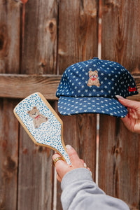 Blue cap with star pattern and teddy bear embroidery held next to a teddy bear brush