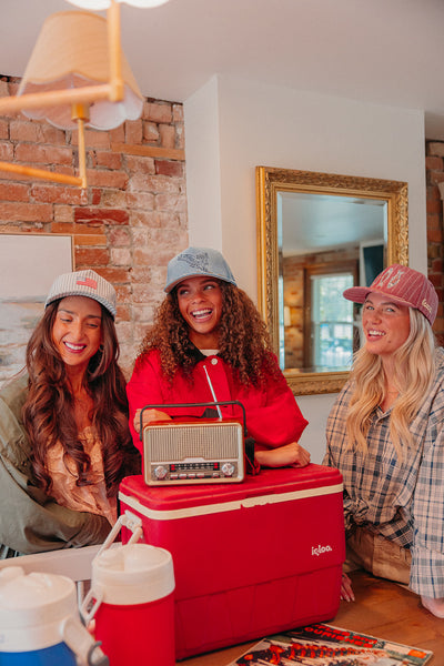Three women sitting around a red cooler with a radio, smiling in a casual indoor setting wearing different hats