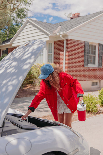 Woman in red jacket and blue cap checking car engine wearing a blue denim trucker hat