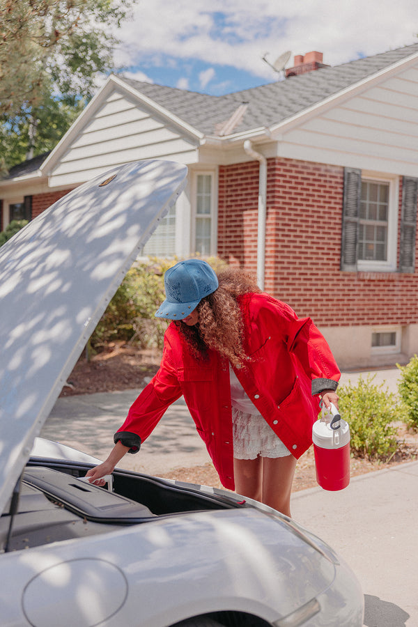 Woman in red jacket and blue cap checking car engine wearing a blue denim trucker hat