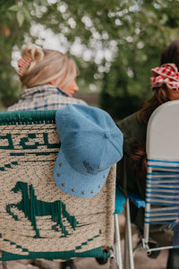 Person sitting on a woven chair with a blue denim trucker hat, surrounded by greenery.