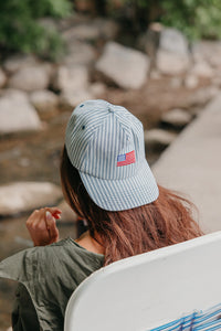 Person wearing a light blue cap with an American flag emblem, sitting by a stream.