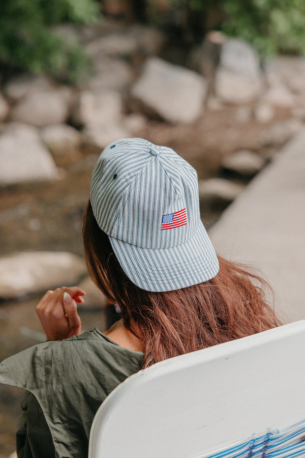 Person wearing a light blue cap with an American flag emblem, sitting by a stream.