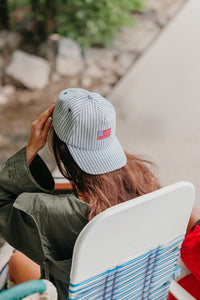 girl wearing a striped baseball cap with an American flag embroidered, sitting by a body of water