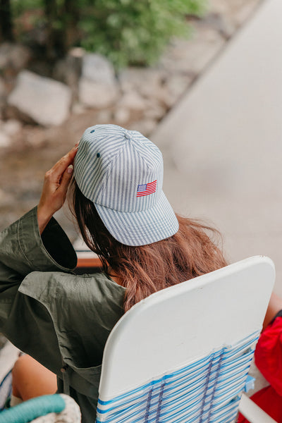 girl wearing a striped baseball cap with an American flag embroidered, sitting by a body of water