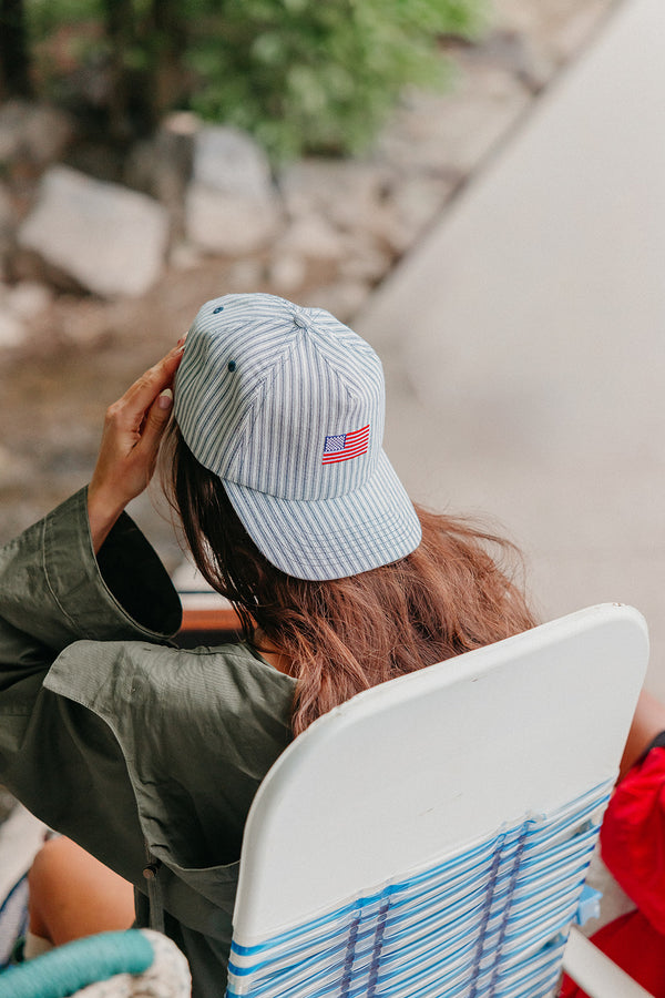 girl wearing a striped baseball cap with an American flag embroidered, sitting by a body of water