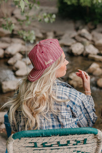 Woman wearing a red cap with kaxi logo on the side, sitting by a stream with rocks and greenery.