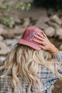 Person wearing a red trucker hat with fish designs, standing outdoors near rocks and water.