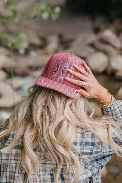Person wearing a red trucker hat with fish designs, standing outdoors near rocks and water.