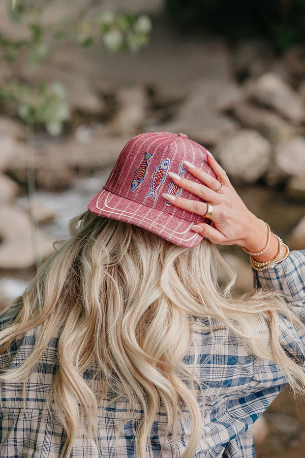 Person wearing a red trucker hat with fish designs, standing outdoors near rocks and water.