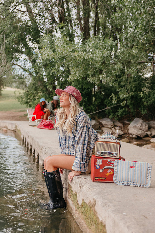 Woman sitting by a river wearing a red trucker hat, with camping gear, including a cooler and boots.
