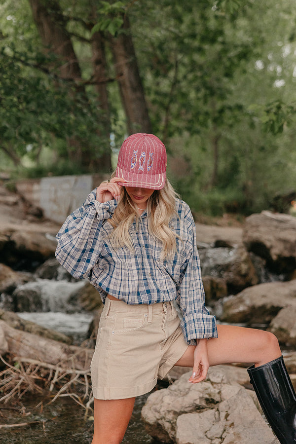 Person wearing a red trucker hat with fish graphic on it, standing on rocks near water and trees.