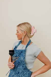 Woman wearing a blue apron holding a black brush against a beige background with pink HAIR GRL claw