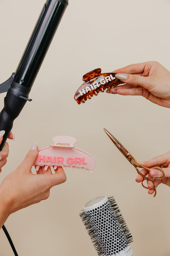 Hands holding hair styling tools with 'HAIR GIRL' branded clips and scissors on a beige background