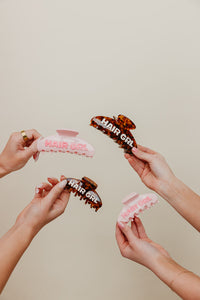 Four hair clips with 'HAIR GIRL' text held by hands against a beige background