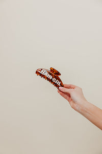 Hand holding a tortoiseshell claw clip with 'HAIR-GRL' text on a beige background