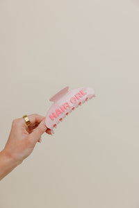 Pink claw clip with 'HAIR GIRL' text held by a hand against a beige background