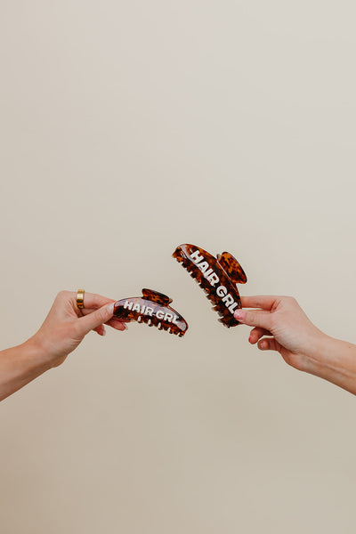 Two hands holding brown hair clips with 'Hair Girl' branding against a beige background