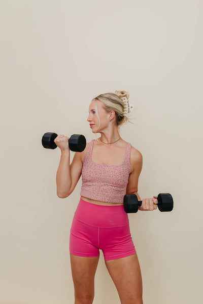 Woman in pink athletic wear lifting dumbbells wearing cream and brown gym girl claw against a beige background